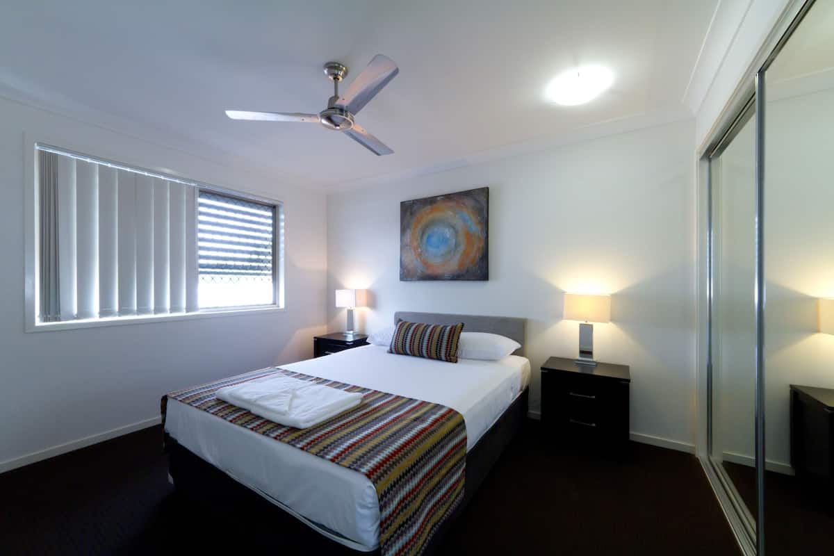 A hotel room with a bed, a ceiling fan, a window with blinds, a painting on the wall, and two bedside tables with lamps.