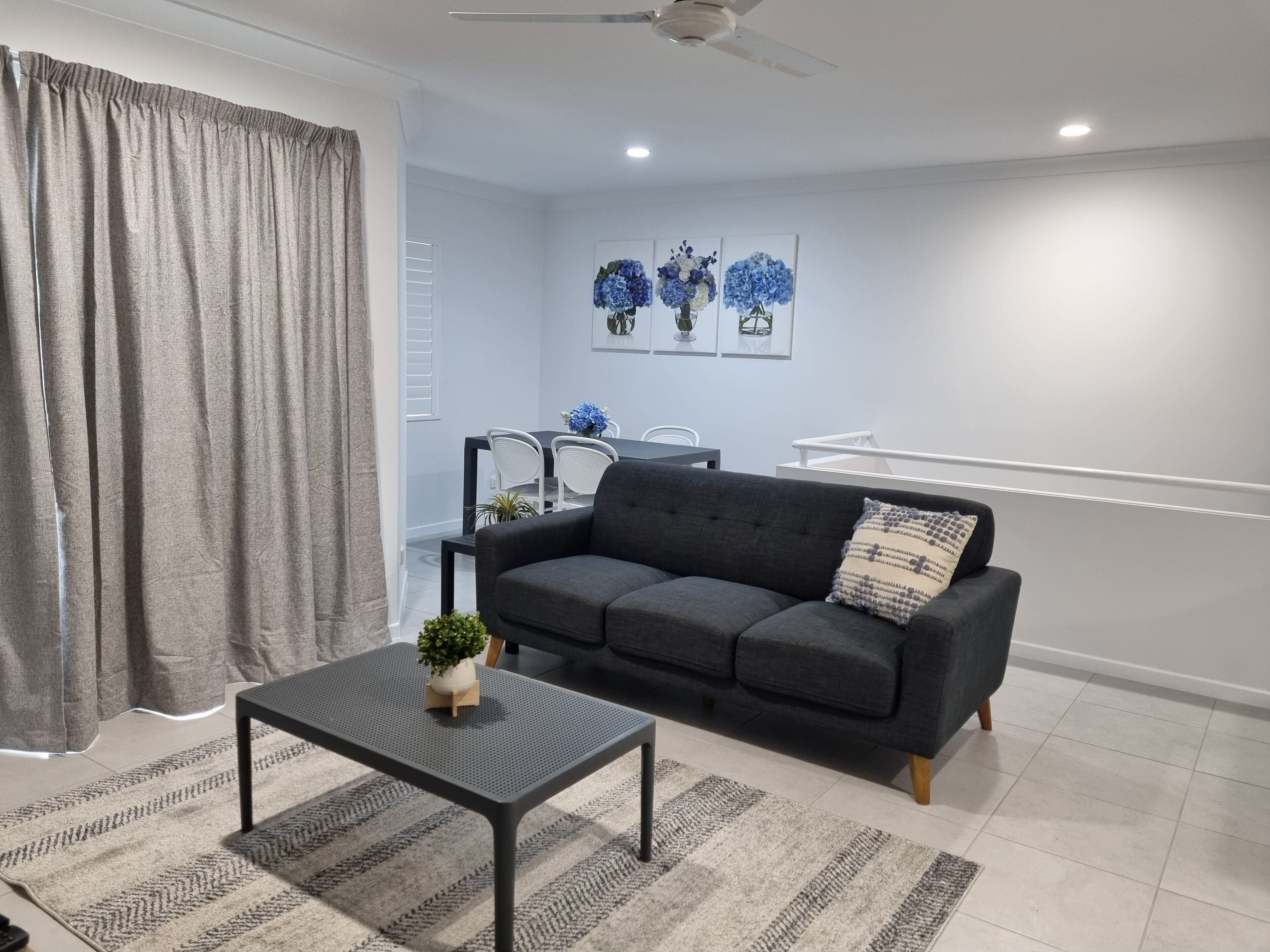A modern living room with a grey sofa, a small table with a plant, and a grey rug. There is a dining area with a table and chairs in the background.