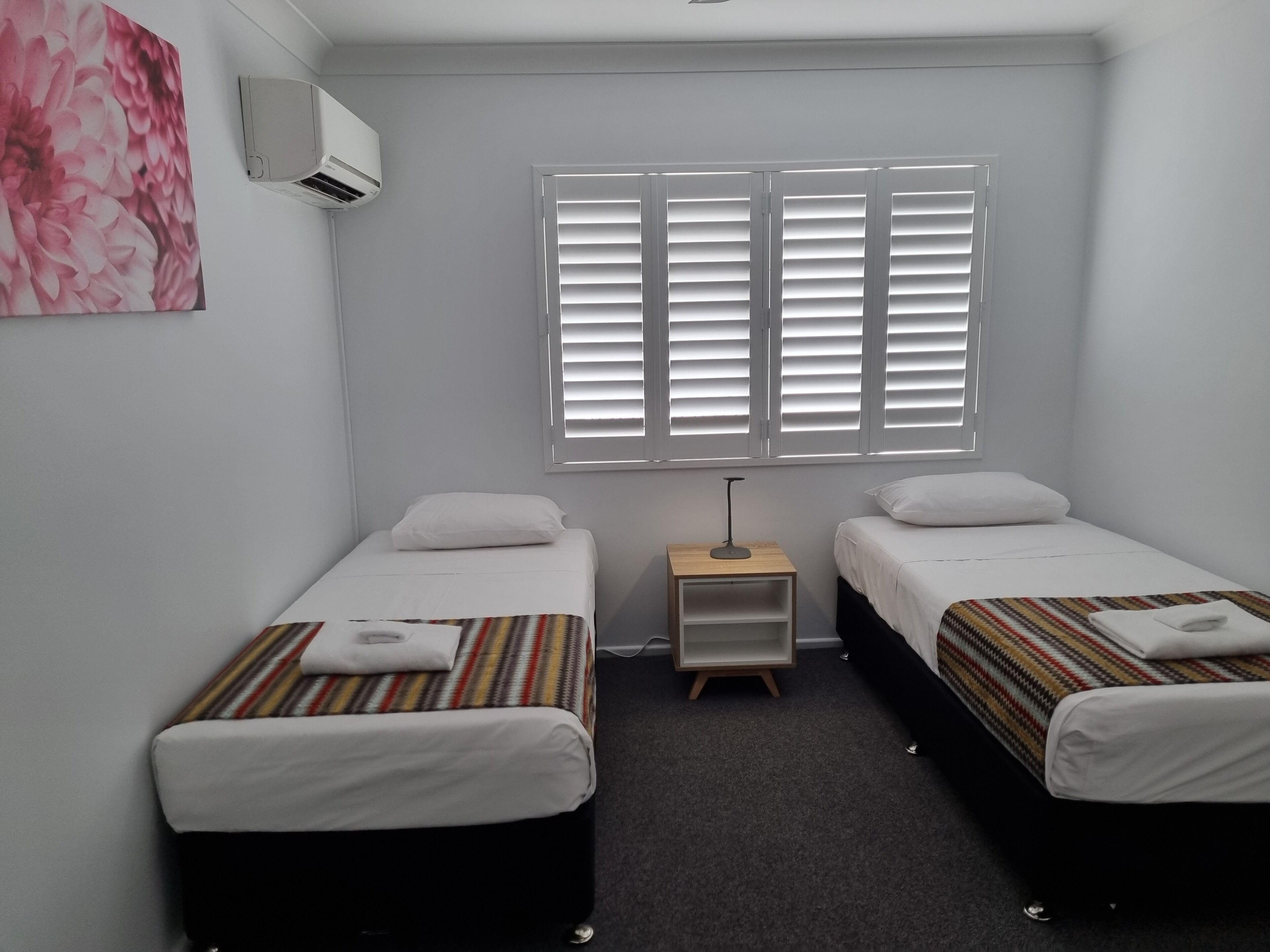 Een kamer met twee eenpersoonsbedden, een klein nachtkastje met een lamp, een raam met zonwering en een airconditioning op de muur.