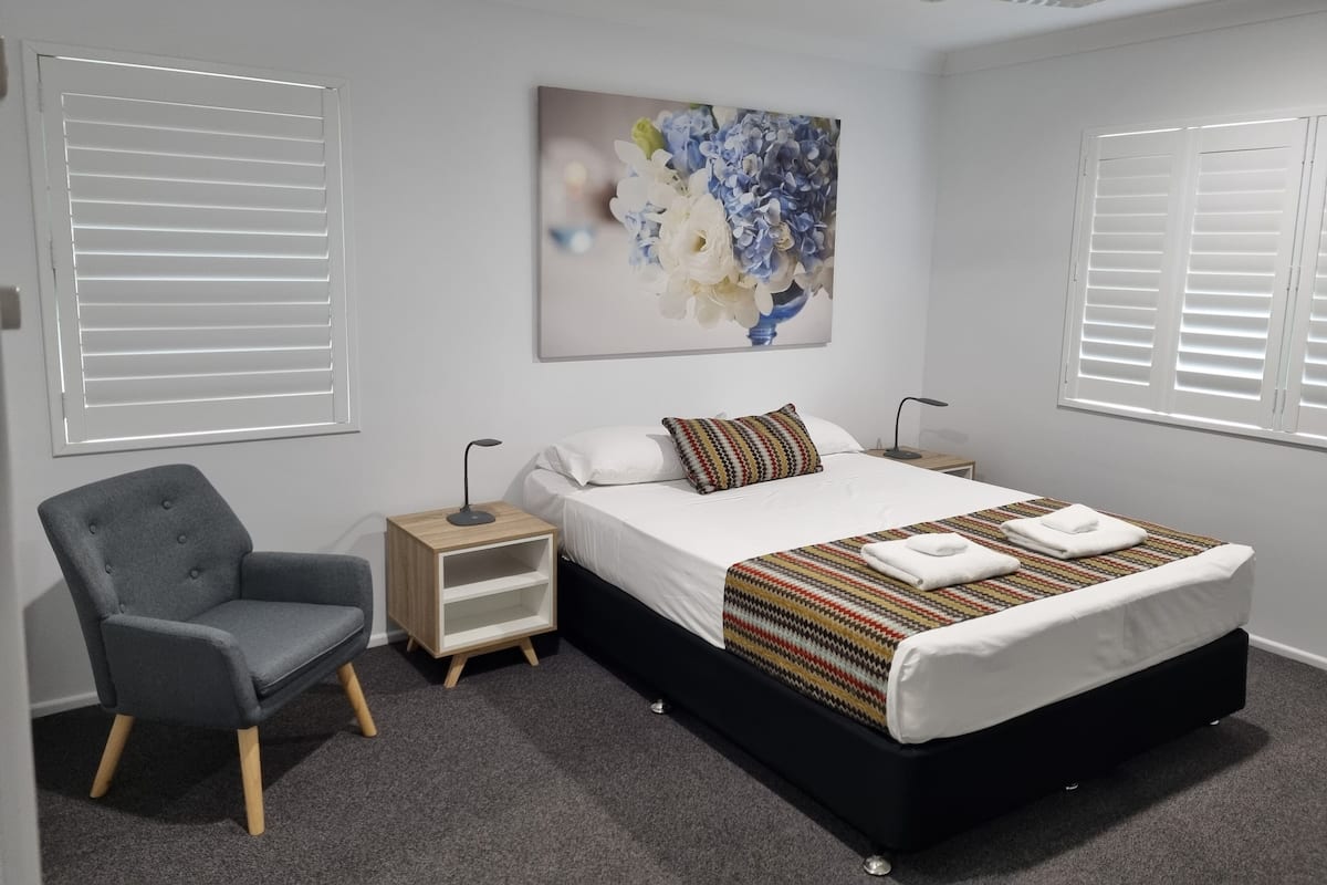 A modern bedroom with a bed, a chair, a nightstand, a ceiling fan, and a wall-mounted artwork.