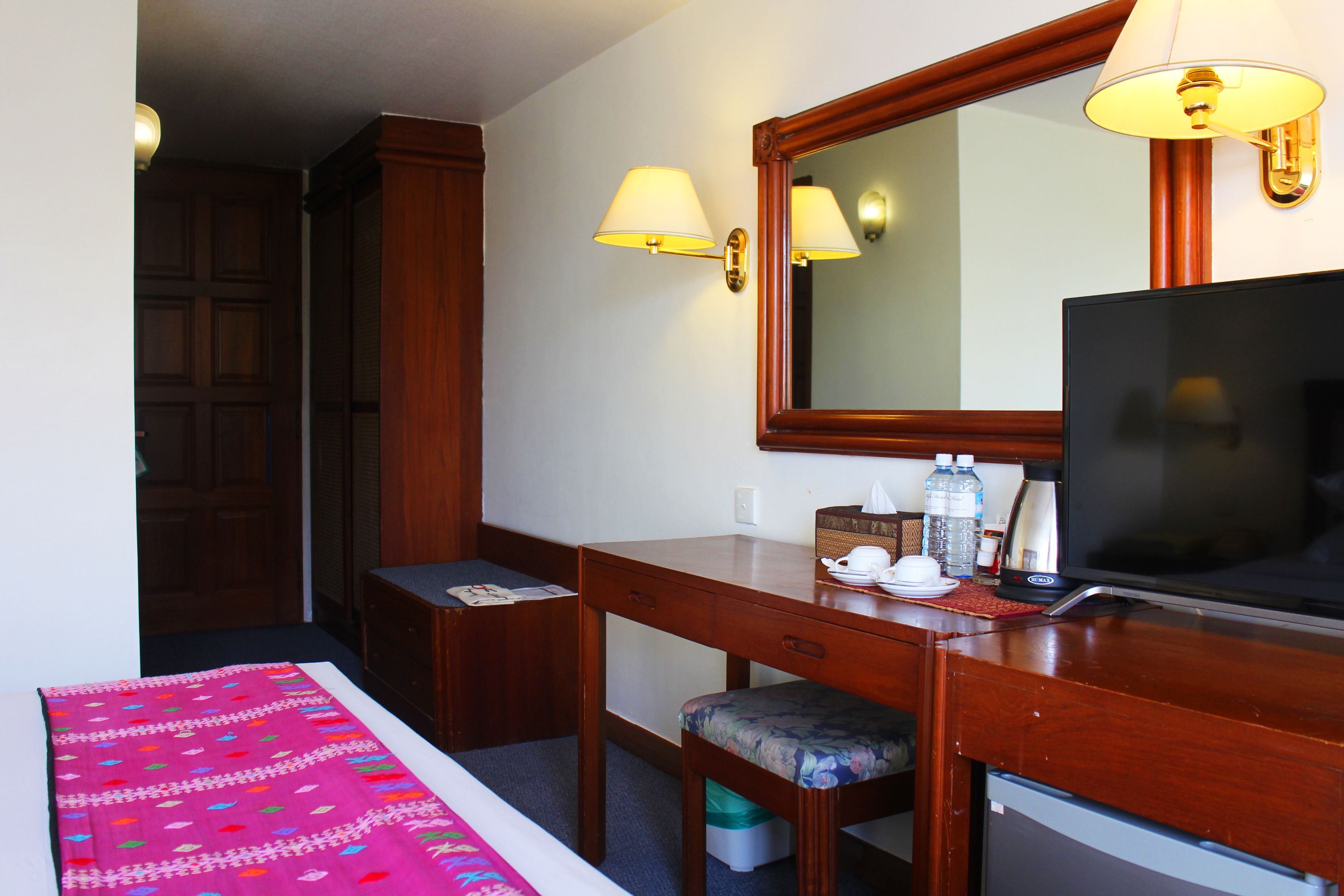A hotel room with a wooden desk, a television, a mirror, and two wall-mounted lamps.