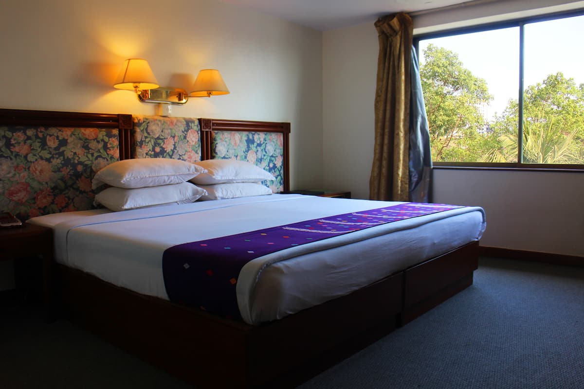 A neatly made bed with a floral headboard, two bedside lamps, and a view of trees through a window.