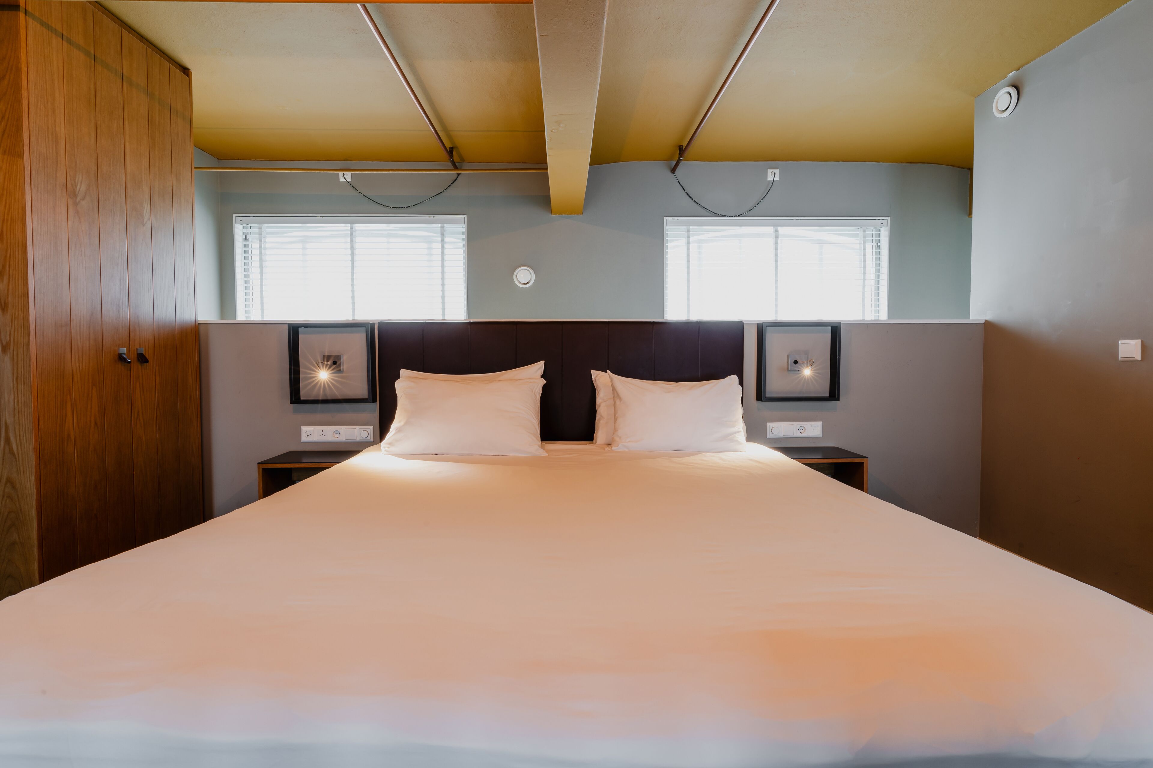 A large bed with white linens, two bedside tables with lamps, wooden wardrobe, and two windows with blinds.