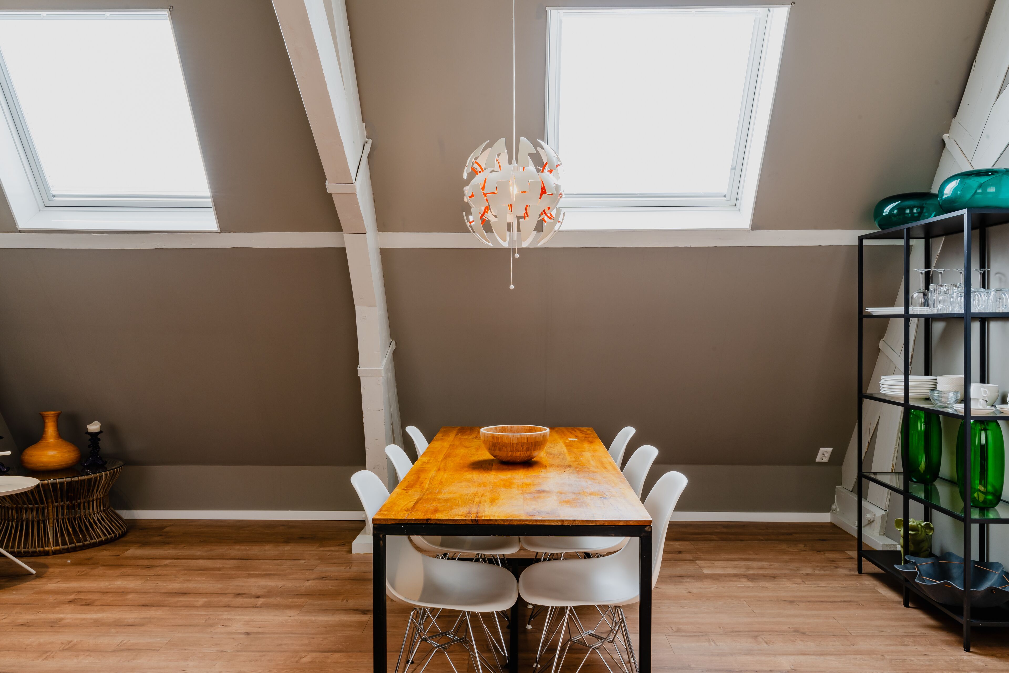 Un coin repas comprenant une table en bois, des chaises blanches, un luminaire suspendu et une étagère où sont rangés des verres.