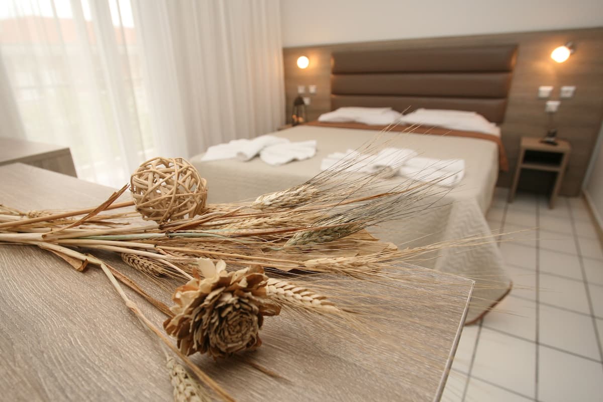 Un letto rifatto con lenzuola bianche e una testiera in legno, un comodino con una lampada e una finestra con tende leggere.