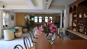 Zona pranzo con un tavolo lungo, sedie e un vaso di fiori. In secondo piano si vedono tavoli e sedie da pranzo, e un'area bar con scaffali pieni di bottiglie.