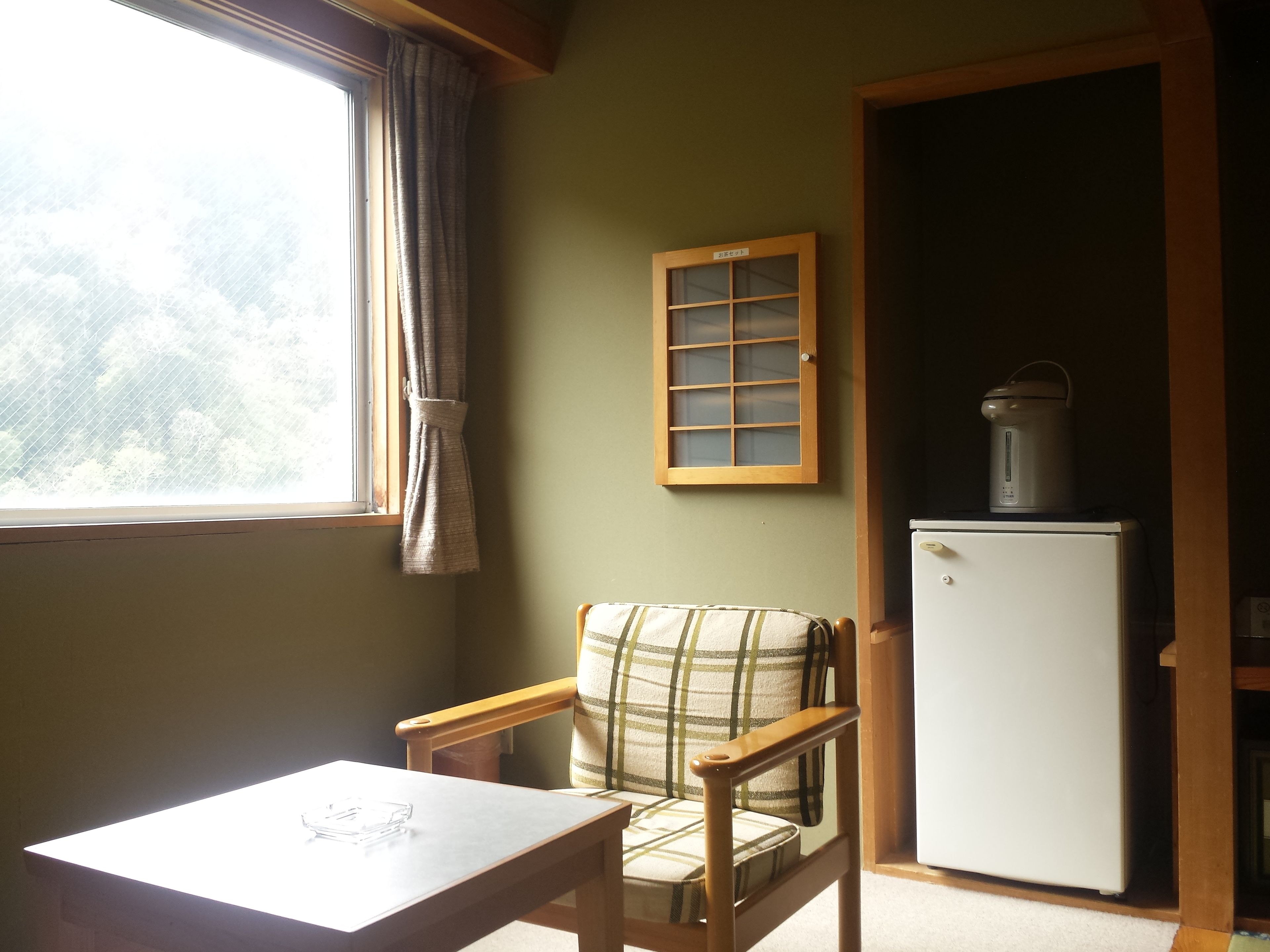 Una habitación pequeña con ventana, una silla, una mesa y un refrigerador.