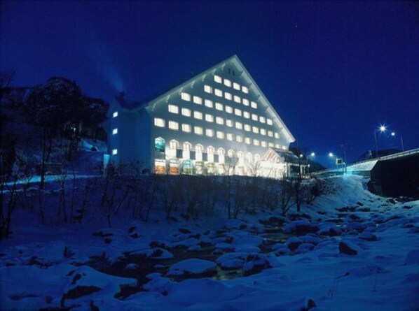 A large, illuminated building with multiple windows and a prominent entrance, surrounded by snow-covered ground and trees.
