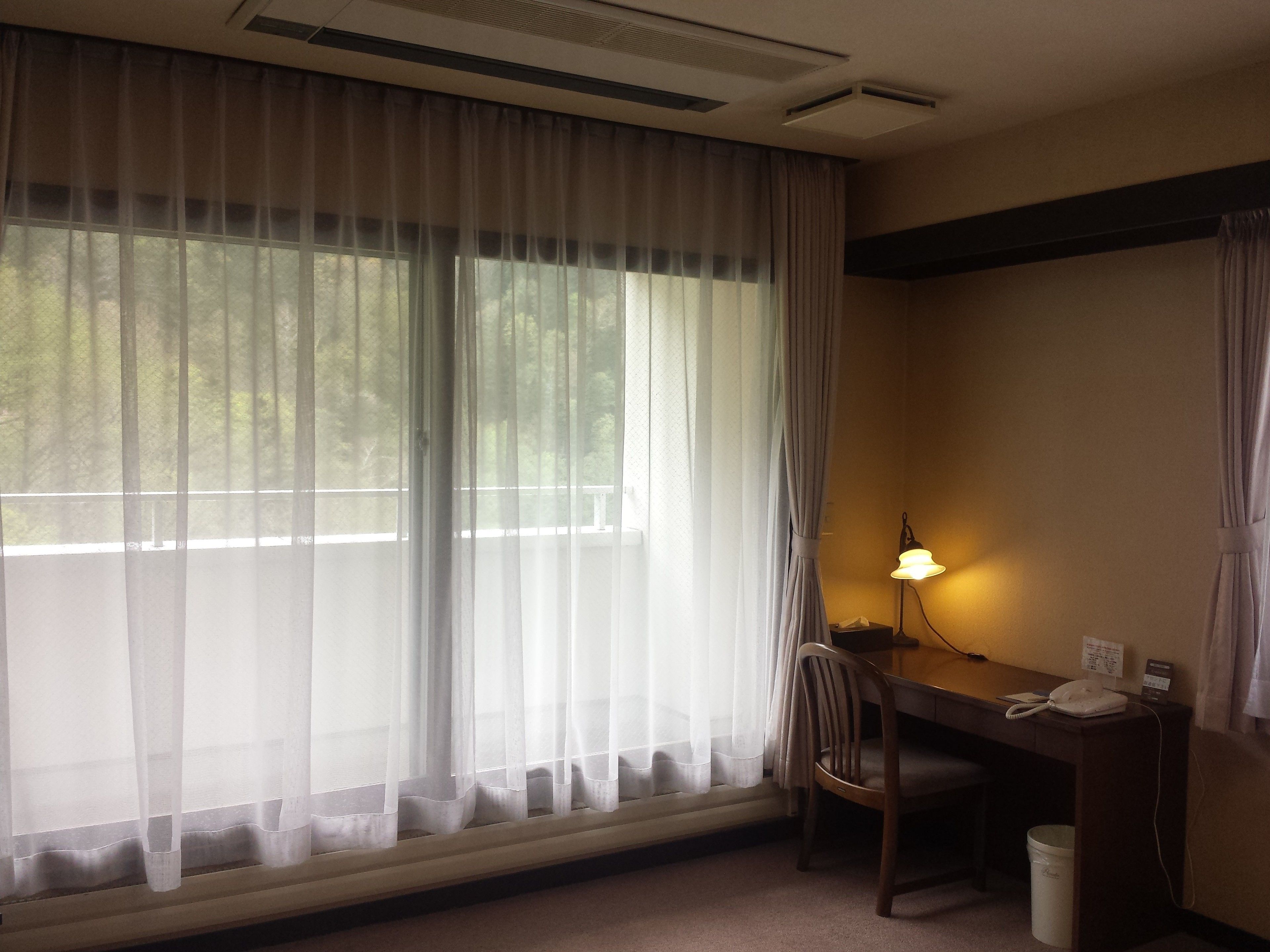 A hotel room with a large window covered by sheer curtains, a desk with a chair, a desk lamp, a telephone, and a waste bin.
