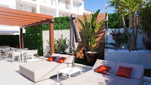 An outdoor seating area with sofas and tables, surrounded by greenery and a white building in the background. - Oasis Sa Tanca Apartments by Typic Hotels (Santa Eulalia del Rio)