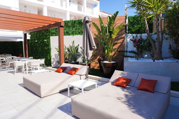 An outdoor seating area with sofas and tables, surrounded by greenery and a white building in the background. - Oasis Sa Tanca Apartments by Typic Hotels (Santa Eulalia del Rio)