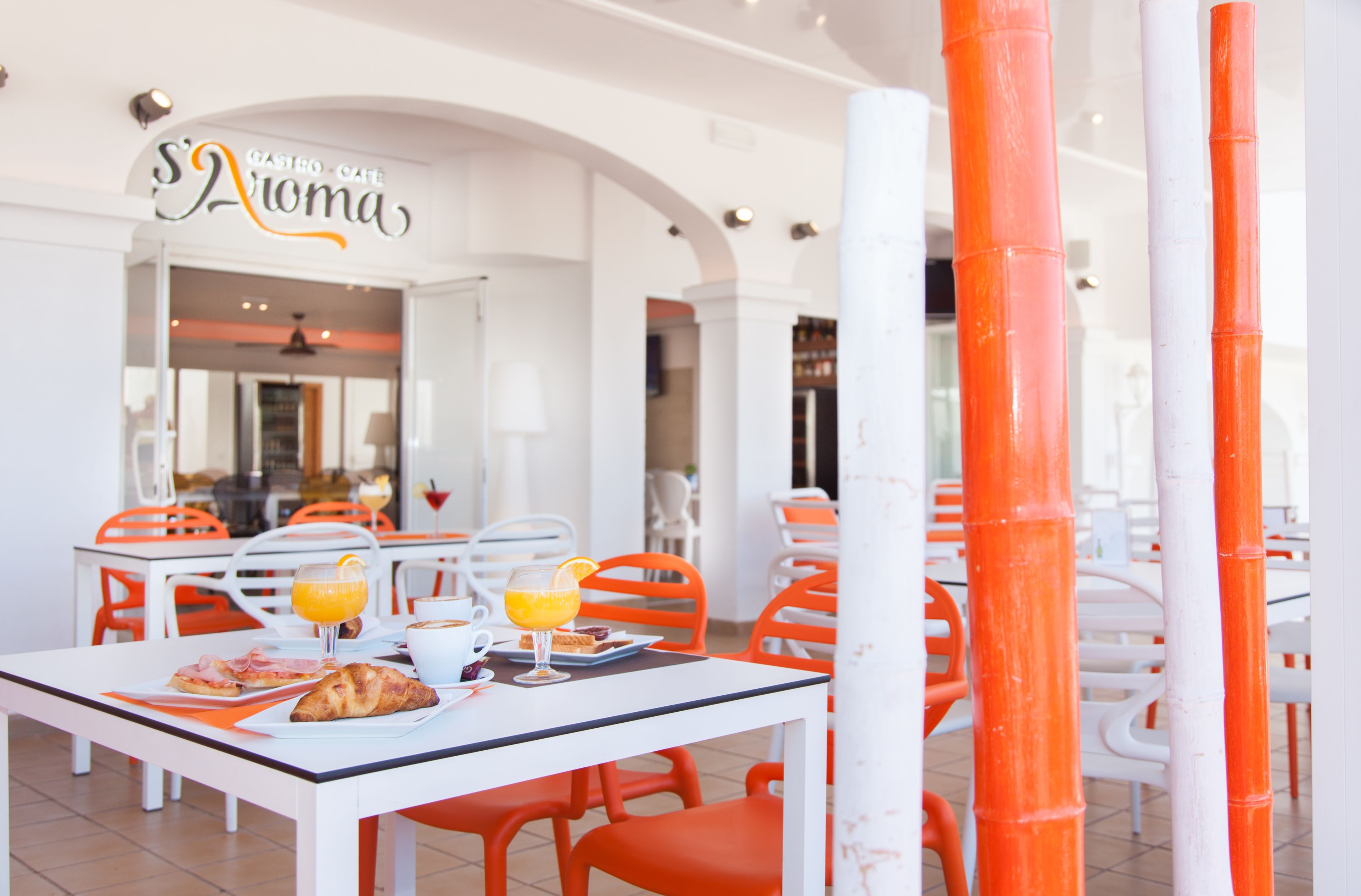 Un comedor con mesas y sillas, un letrero que dice "Gastro-Cafe S'Atoma" y un vaso de zumo de naranja sobre una mesa.