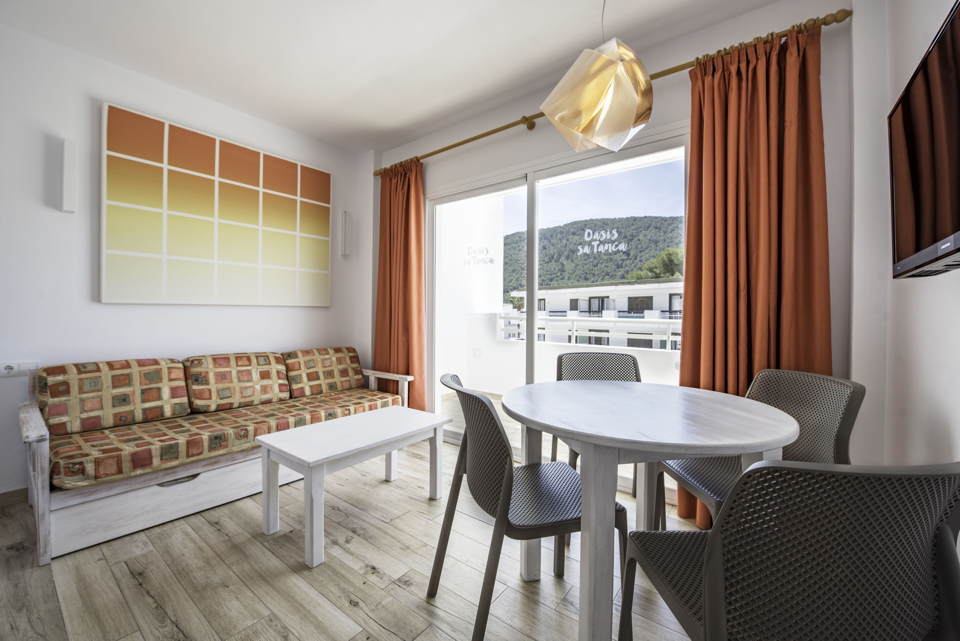 Un salon moderne avec un canapé, une table à manger et des chaises. Une grande fenêtre, ornée de rideaux, offre une vue sur un immeuble et des collines à l’extérieur.