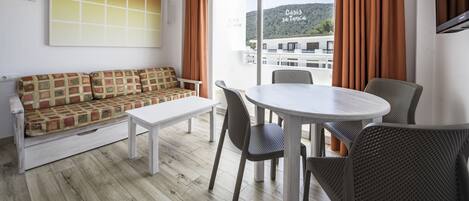 Un salon moderne avec un canapé, une table à manger et des chaises. Une grande fenêtre, ornée de rideaux, offre une vue sur un immeuble et des collines à l’extérieur.