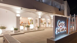 A hotel entrance with a modern design, featuring a large illuminated sign, arched entryways, and potted plants. - Oasis Sa Tanca Apartments by Typic Hotels (Santa Eulalia del Rio)