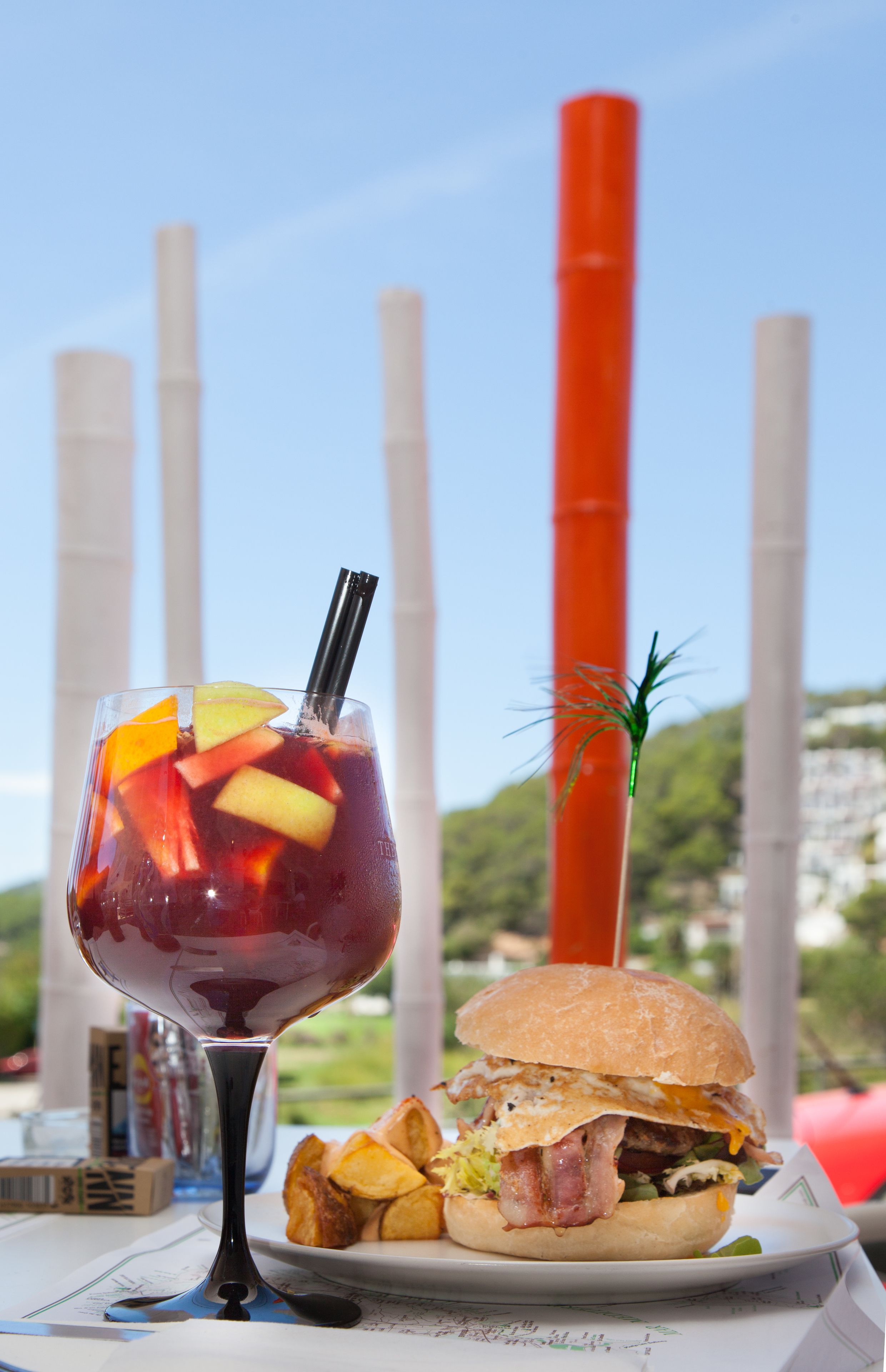Un verre d’une boisson rouge aux fruits, agrémenté d’une paille, un hamburger au bacon et des frites de pommes de terre, le tout sur une table d’où l’on peut admirer de grands bâtiments en arrière-plan.
