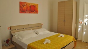 A hotel room with a bed, a wooden wardrobe, a painting of flowers on the wall, and a nightstand with a lamp. - Hotel Santa Costanza (San Vincenzo)