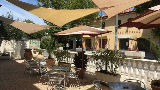 An outdoor patio with metal tables and chairs, shaded by large umbrellas, and surrounded by potted plants and a building with a sign reading "Hotel Santa Costanza". - Hotel Santa Costanza (San Vincenzo)