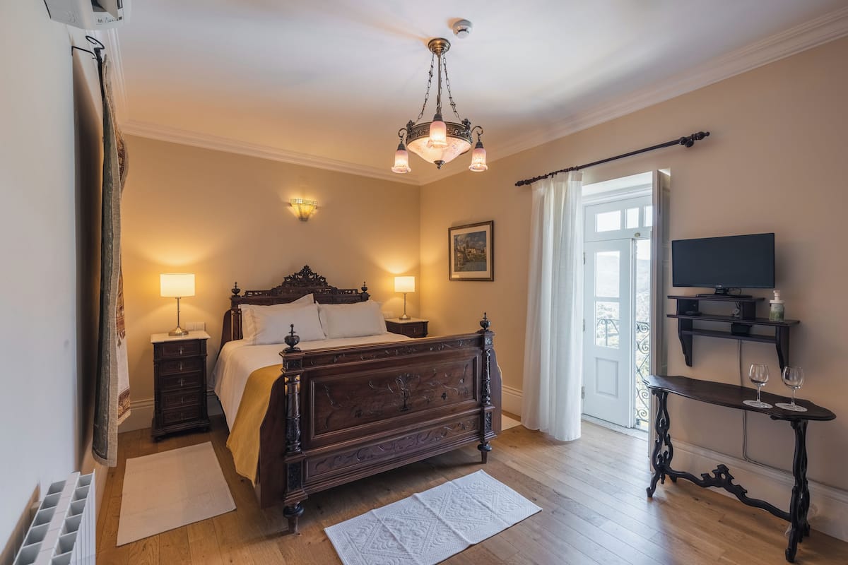 A bedroom with a wooden bed, bedside lamps, a television on a stand, and a view of the outdoors.