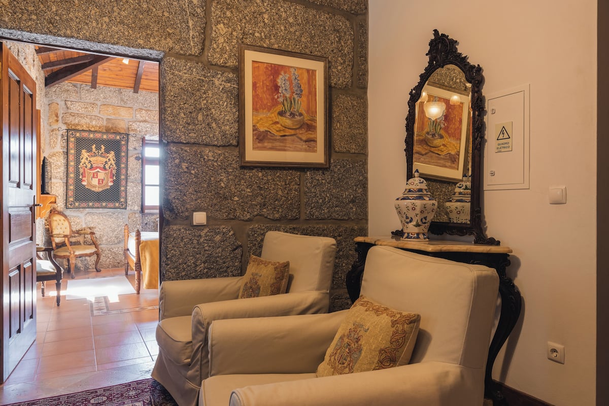 A room with two beige armchairs, a stone wall, a framed painting, and a mirror with a decorative frame.