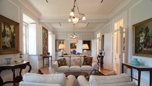 Un salón espacioso con un sofá blanco grande, una mesa de centro y una araña. Hay pinturas en las paredes y un comedor al fondo.