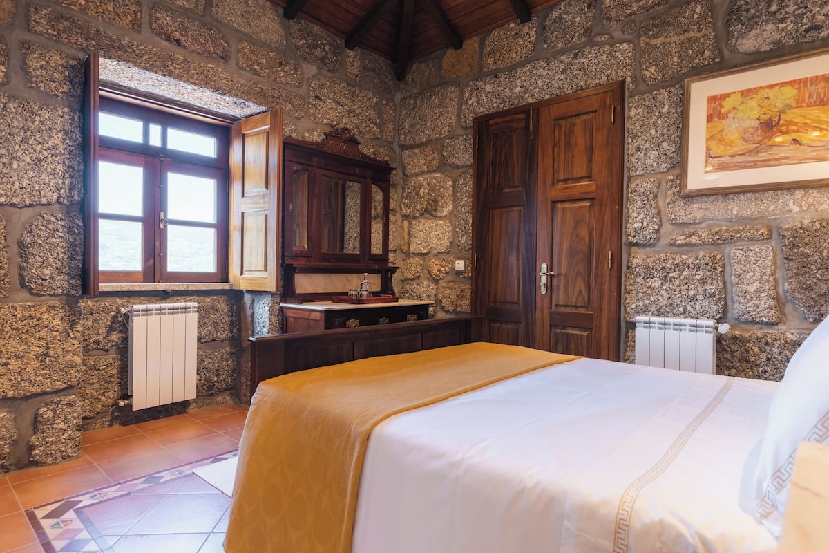 A stone-walled room with a bed, wooden wardrobe, and a window with a view.