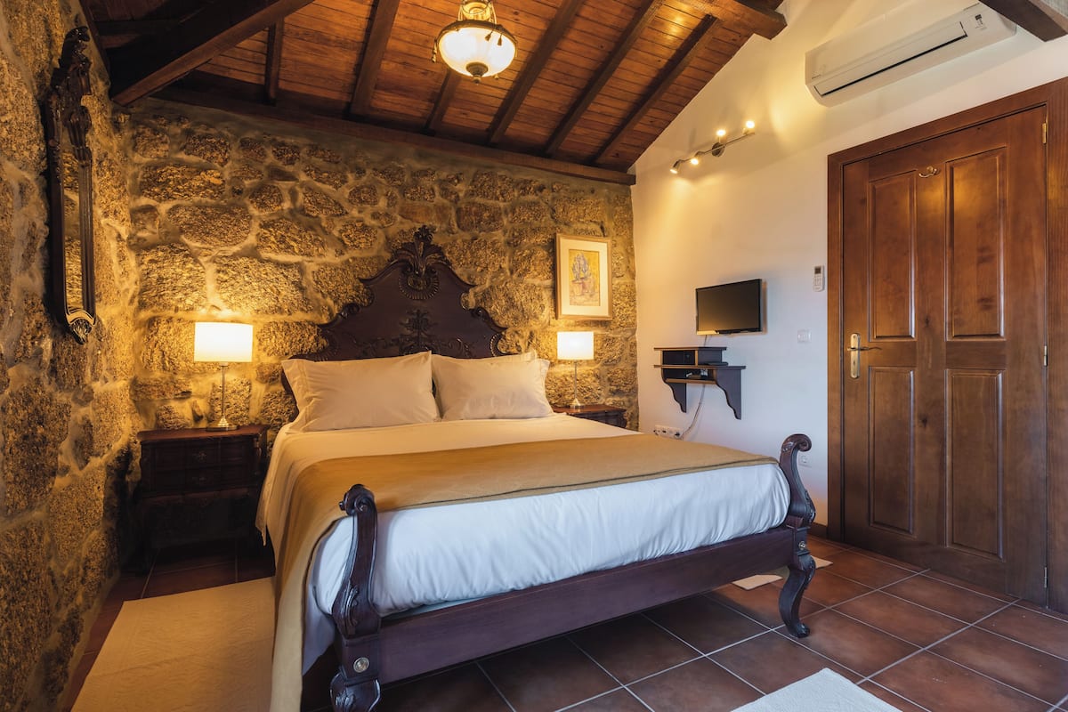 A bedroom with a stone wall, a wooden bed, two bedside lamps, a mounted TV, and a wooden door.