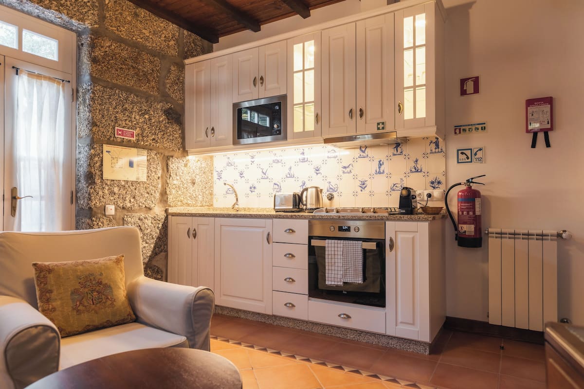 A kitchen with white cabinets, a microwave, an oven, and a fire extinguisher. There is a chair with a cushion and a window with curtains.
