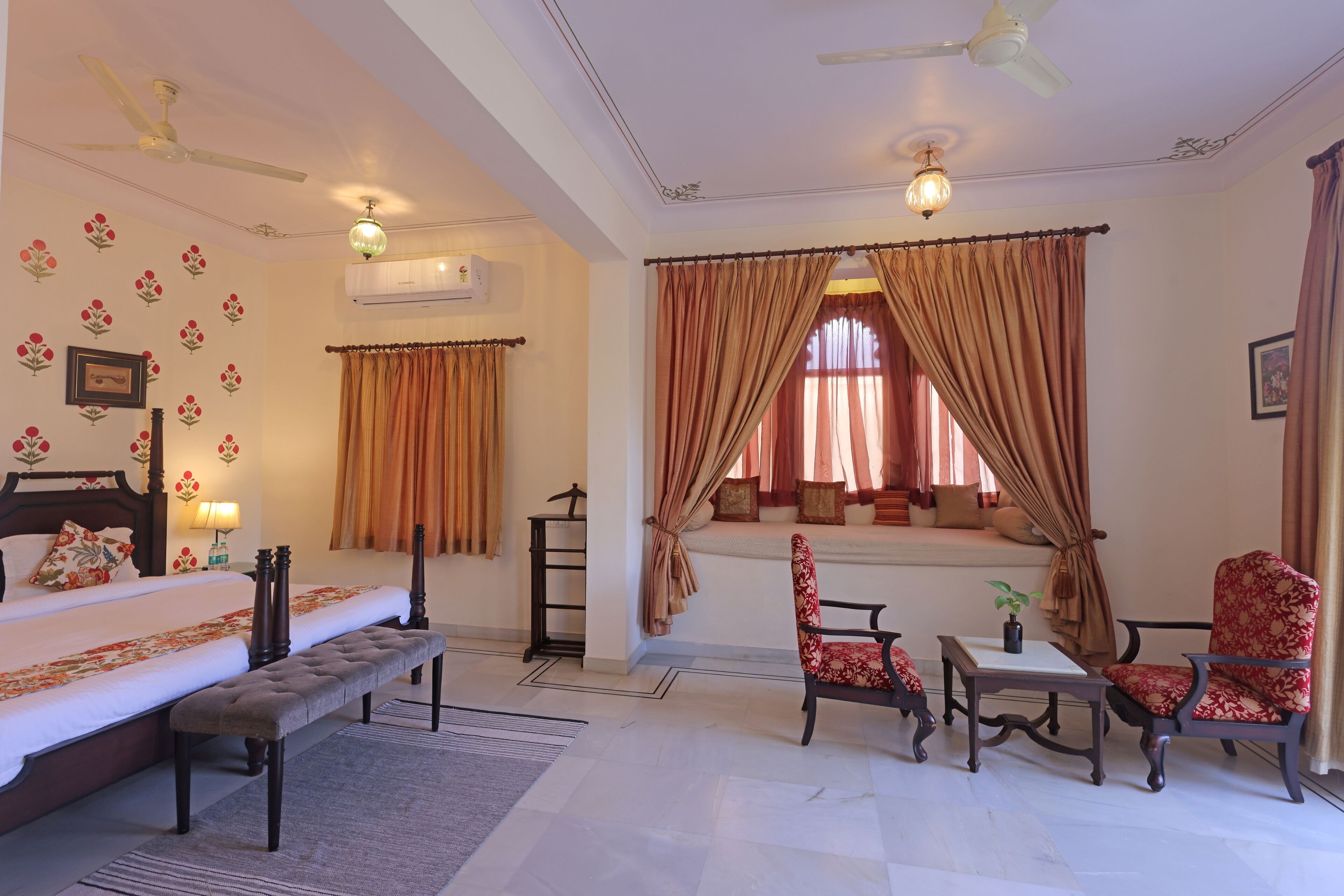 Une chambre d’hôtel comprenant un lit, un banc, deux chaises, une table, une fenêtre avec des rideaux, un ventilateur de plafond et un climatiseur fixé au mur.