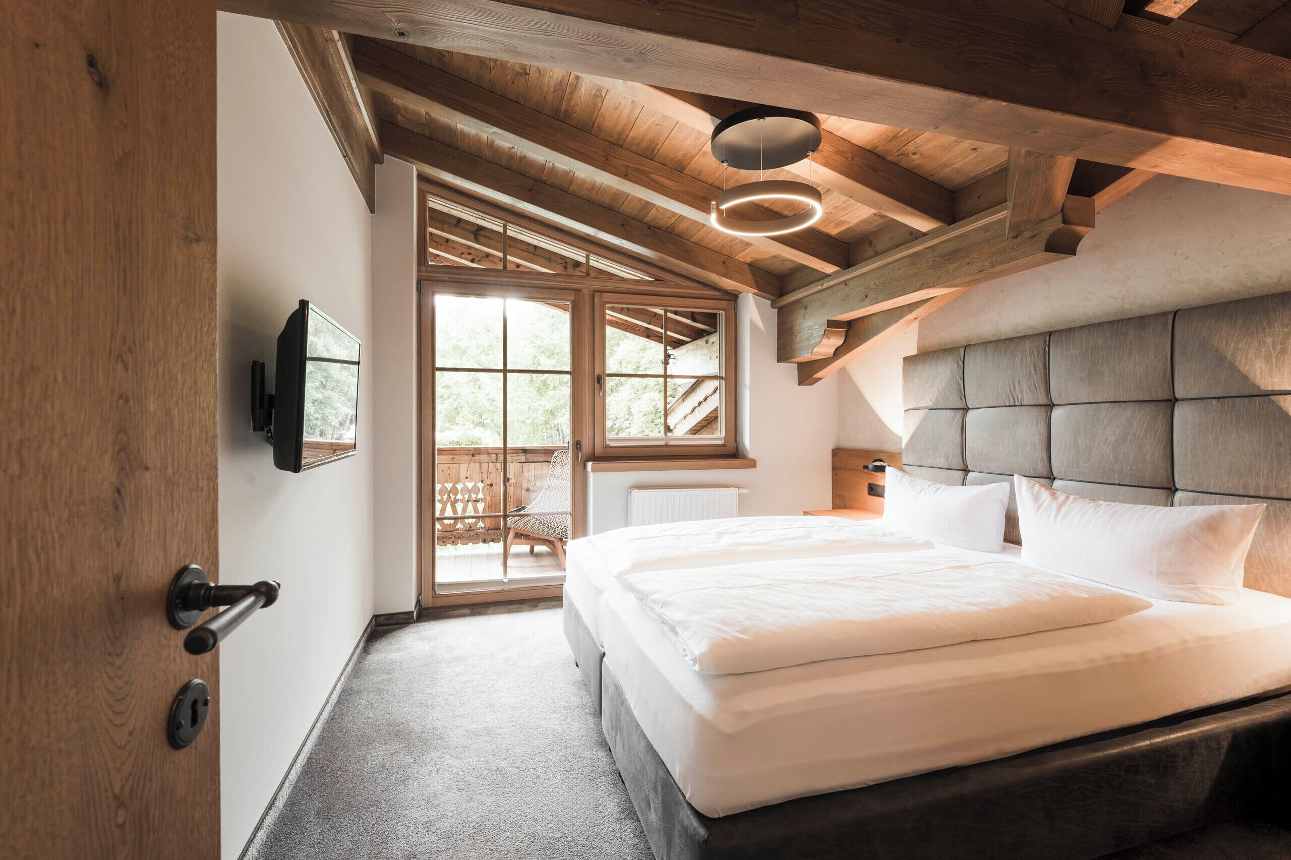 Ein Schlafzimmer mit einem großen Bett, Holzbalkendecke, einem an der Wand montierten Fernseher und einer Glastür zum Außenbereich.