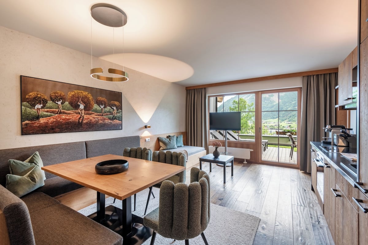 Un salón moderno con una mesa de comedor de madera, asientos tapizados en gris, un cuadro grande y vistas al exterior a través de un ventanal corredero.