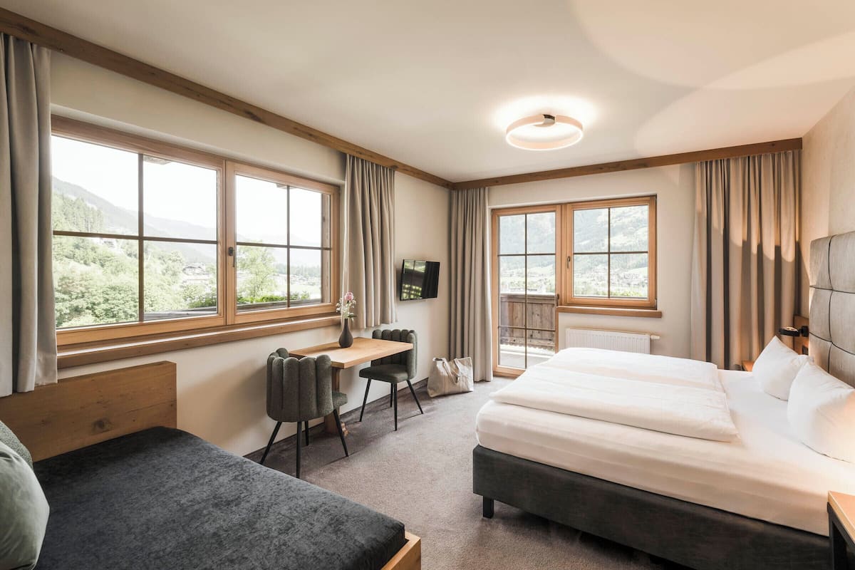 Habitación de hotel con cama, escritorio y silla. Amplias ventanas con cortinas, un televisor de pantalla plana en la pared y un radiador debajo de las ventanas.