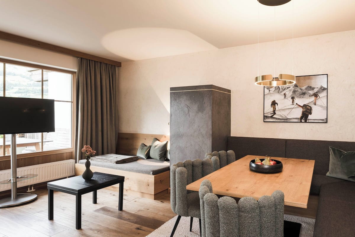Una sala moderna con una mesa de comedor de madera, sillas tapizadas de gris, un sofá, un televisor y un cuadro con esquiadores en la pared.