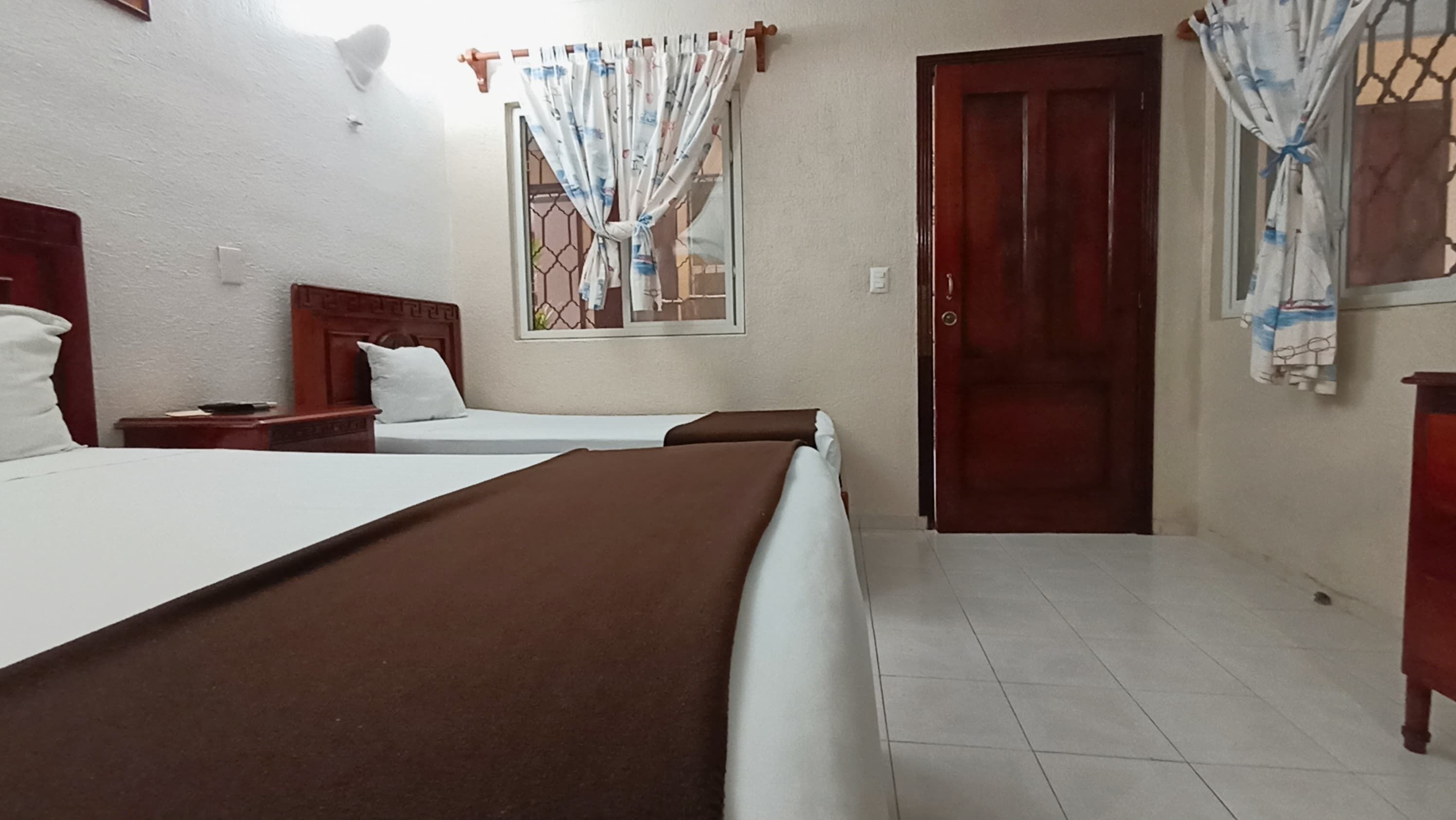 Habitación de hotel con dos camas, cabecero de madera, ventana con cortinas, puerta y suelo de baldosas.