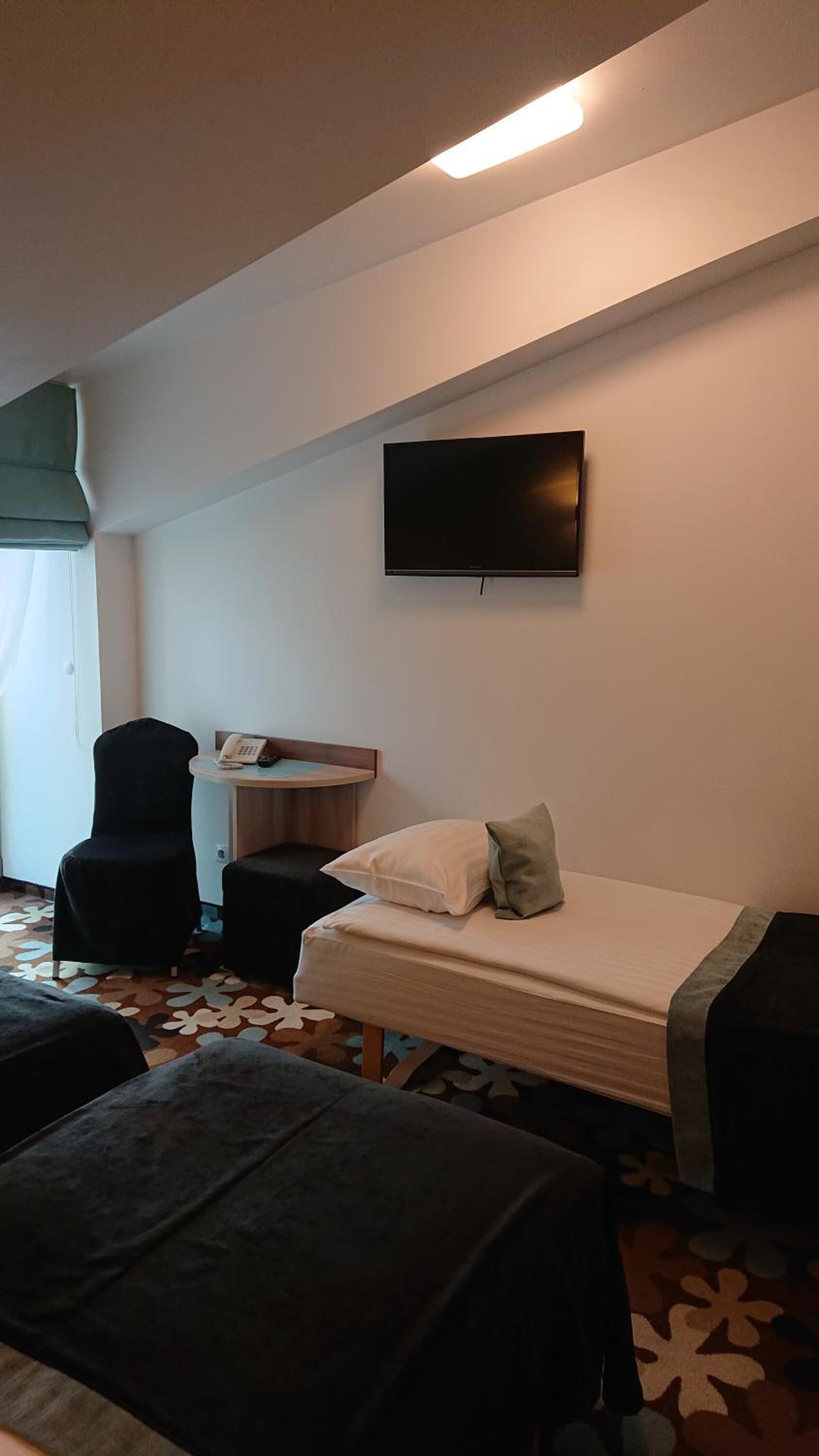 Ein Hotelzimmer mit Bett, Schreibtisch mit Telefon, Stuhl, Wandfernseher und einem gemusterten Teppich.