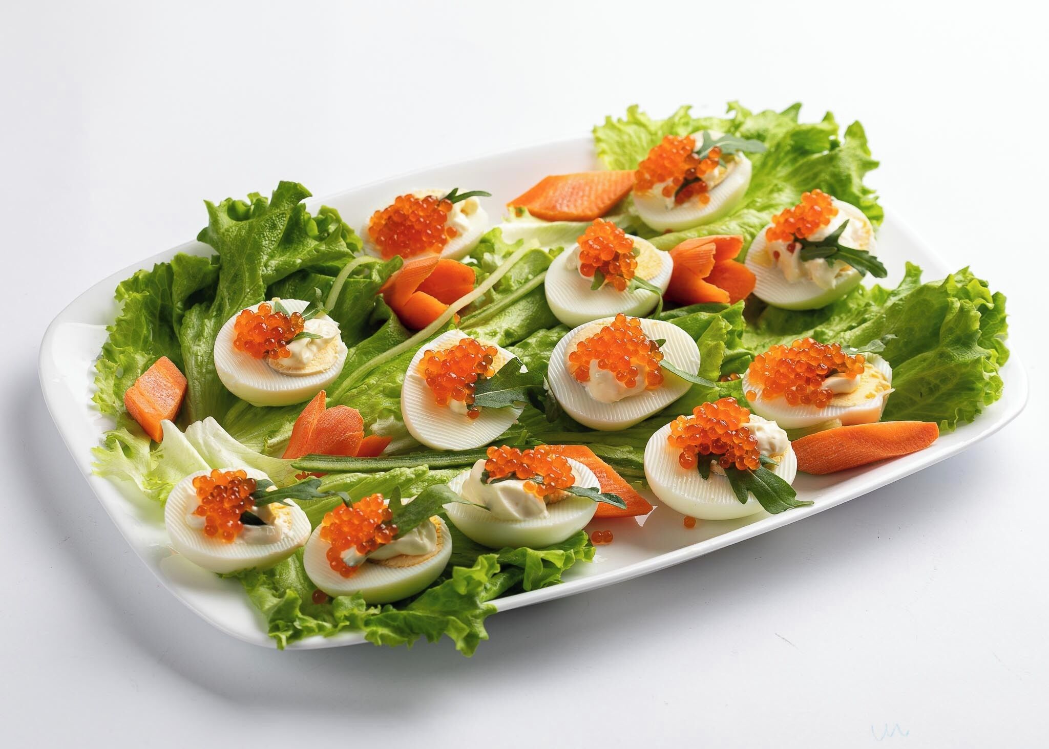 A white rectangular plate with deviled eggs garnished with red caviar, surrounded by lettuce and carrot slices.