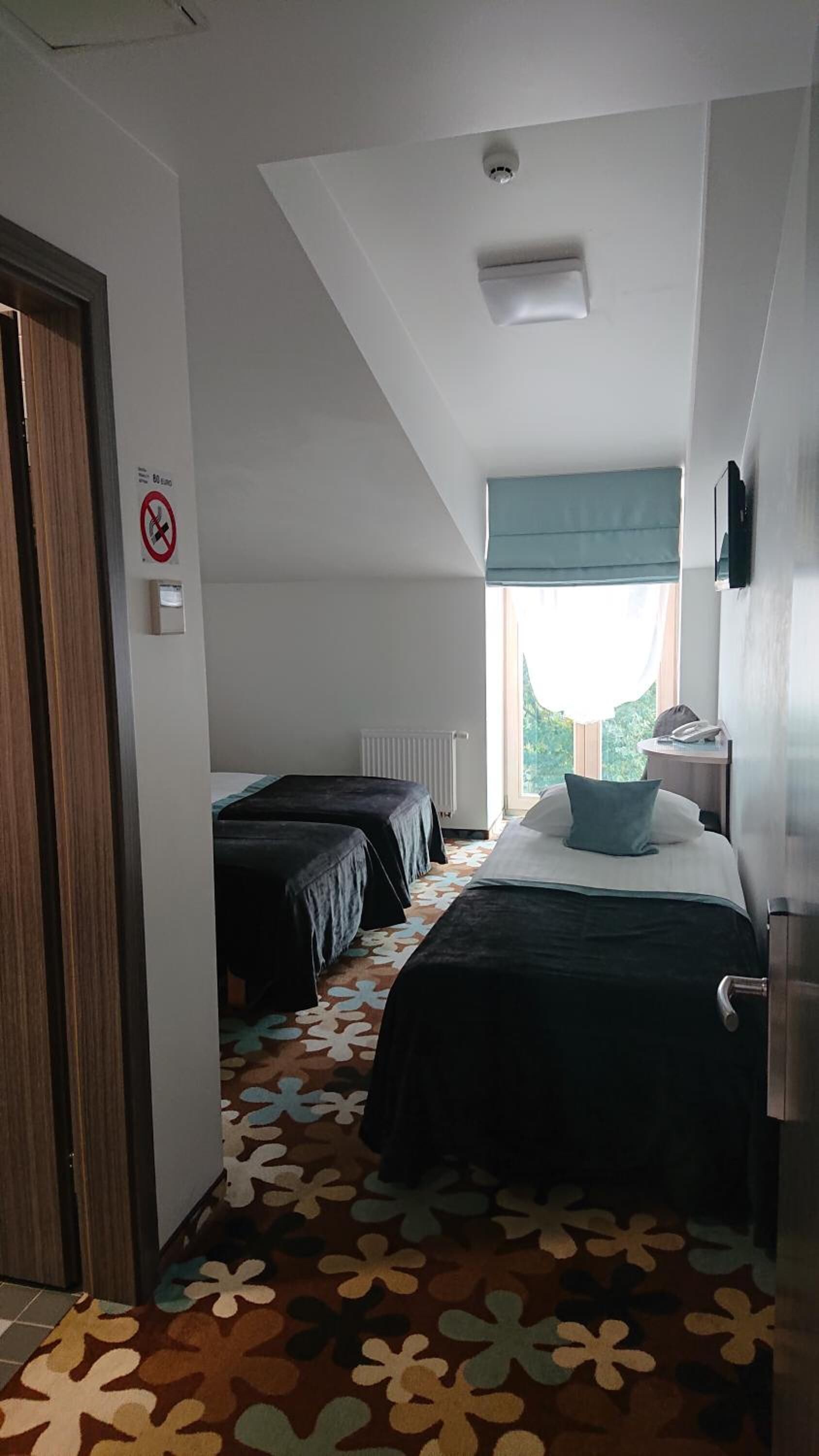 Habitación de hotel con dos camas, un radiador, una ventana con cortinas, una puerta y un cartel de no fumar.