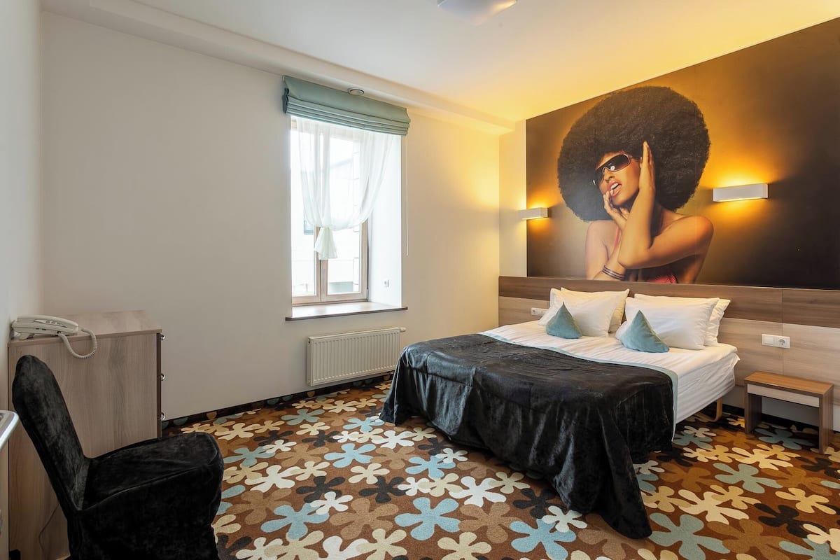 Ein Hotelzimmer mit Bett, Schreibtisch, Stuhl, einem Fenster mit Vorhängen und einer Wandleuchte.