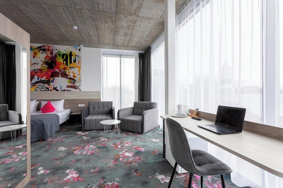 Ein modernes Hotelzimmer mit einem großen Fenster, einem Bett mit einem roten Kissen, zwei grauen Sesseln, einem kleinen runden Tisch und einem Schreibtisch mit einem Laptop.