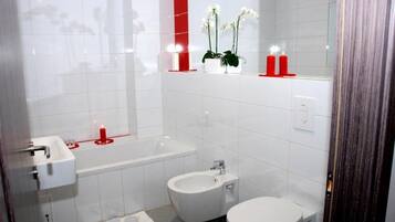 Um banheiro moderno com pia branca, banheira, bidê e vaso sanitário. Há velas vermelhas e orquídeas brancas no lavatório.