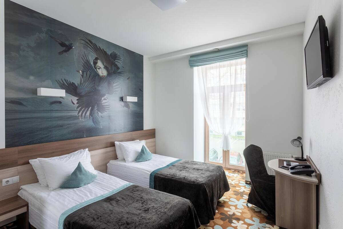 Ein Hotelzimmer mit zwei Betten, einem großen Gemälde an der Wand, einem Schreibtisch mit Telefon und einem fenster mit Vorhängen.