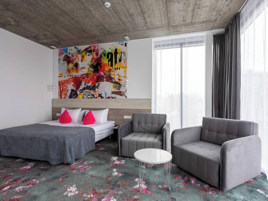Una habitación de hotel moderna con un cuadro abstracto grande, un sofá gris, una mesita redonda pequeña y una cama con ropa de cama gris y almohadas rosas.