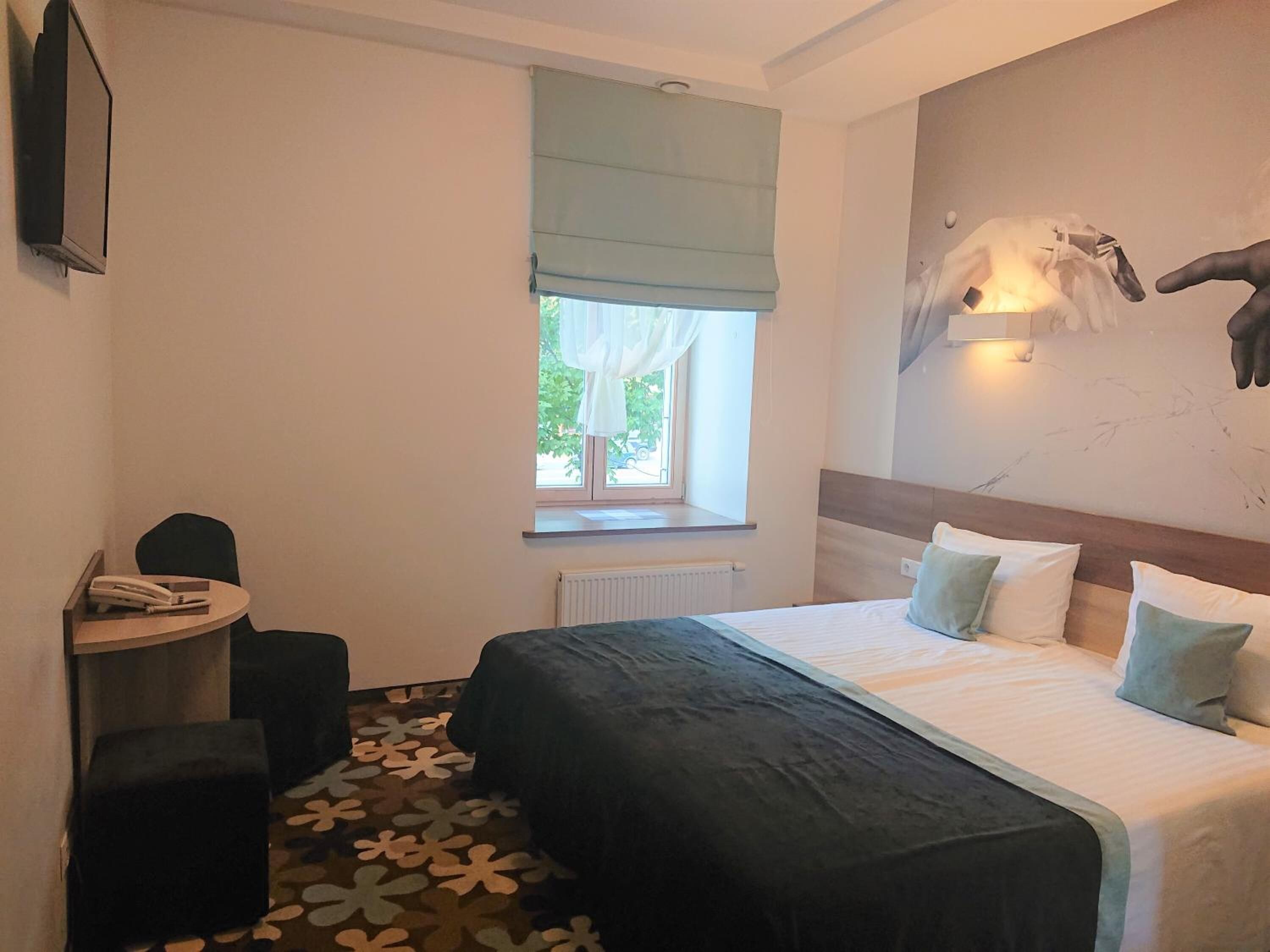 Un dormitorio con cama, una mesita, una silla, un radiador y una ventana con vistas al exterior.