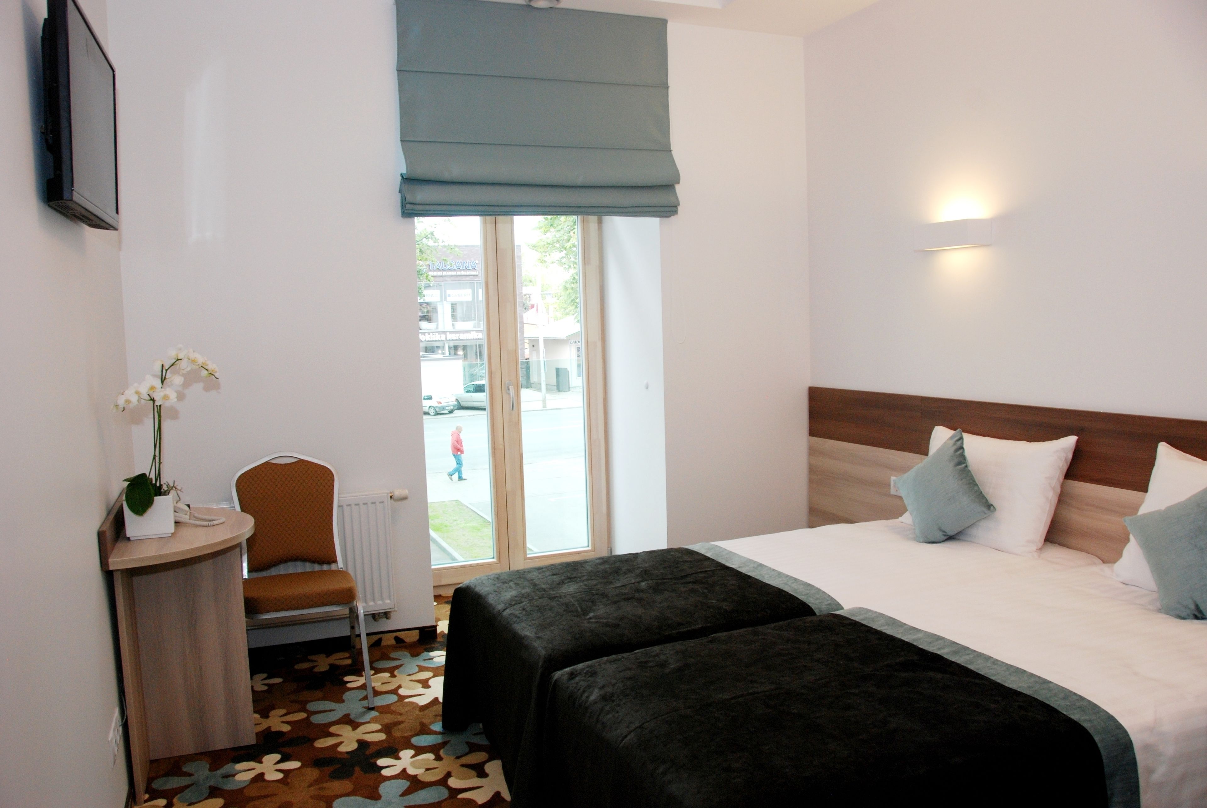 A hotel room with a bed, two bedside tables, a chair, a potted plant, and a window with a view of the street.