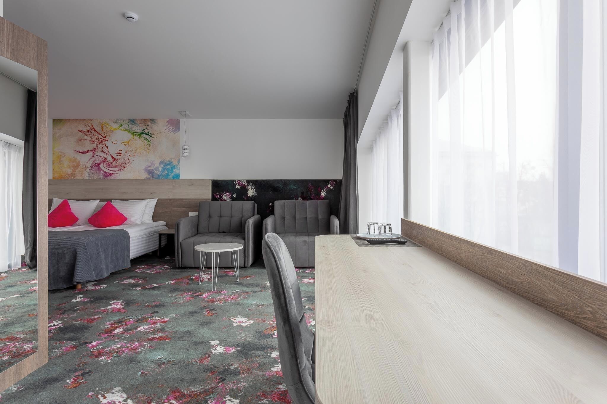 A modern hotel room with a bed, two armchairs, a small table, and a large window.