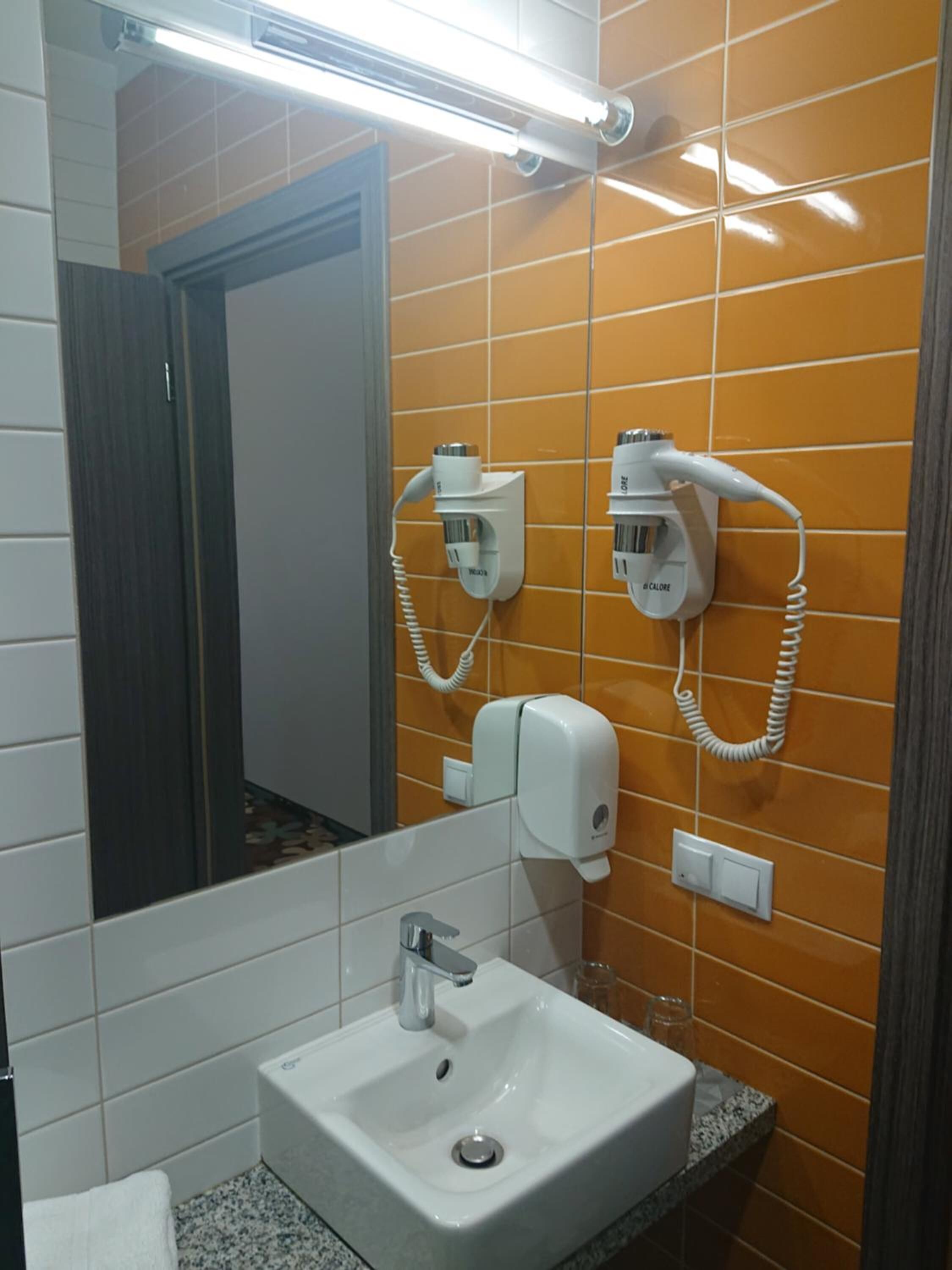 Ein Badezimmer mit einem weißen Waschbecken, zwei an der Wand montierten Haartrocknern, einem Spiegel und einer Leuchte darüber.