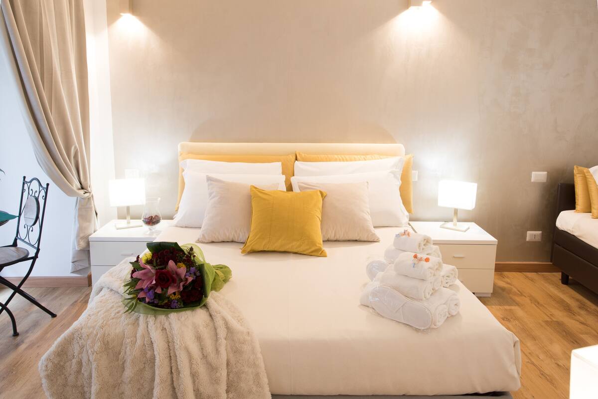 A hotel room with a large bed, two bedside tables with lamps, a chair, and a small table with towels and a bouquet of flowers.