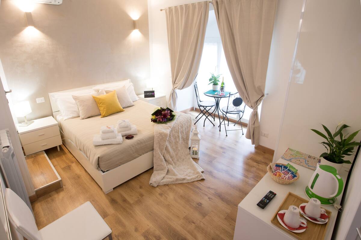 A hotel room with a bed, bedside table, chair, and a small dining area with a table and chairs.