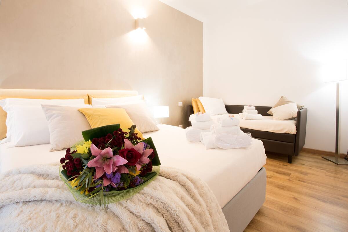 A hotel room with a large bed, a bedside table with a lamp, and a bouquet of flowers on the bed.