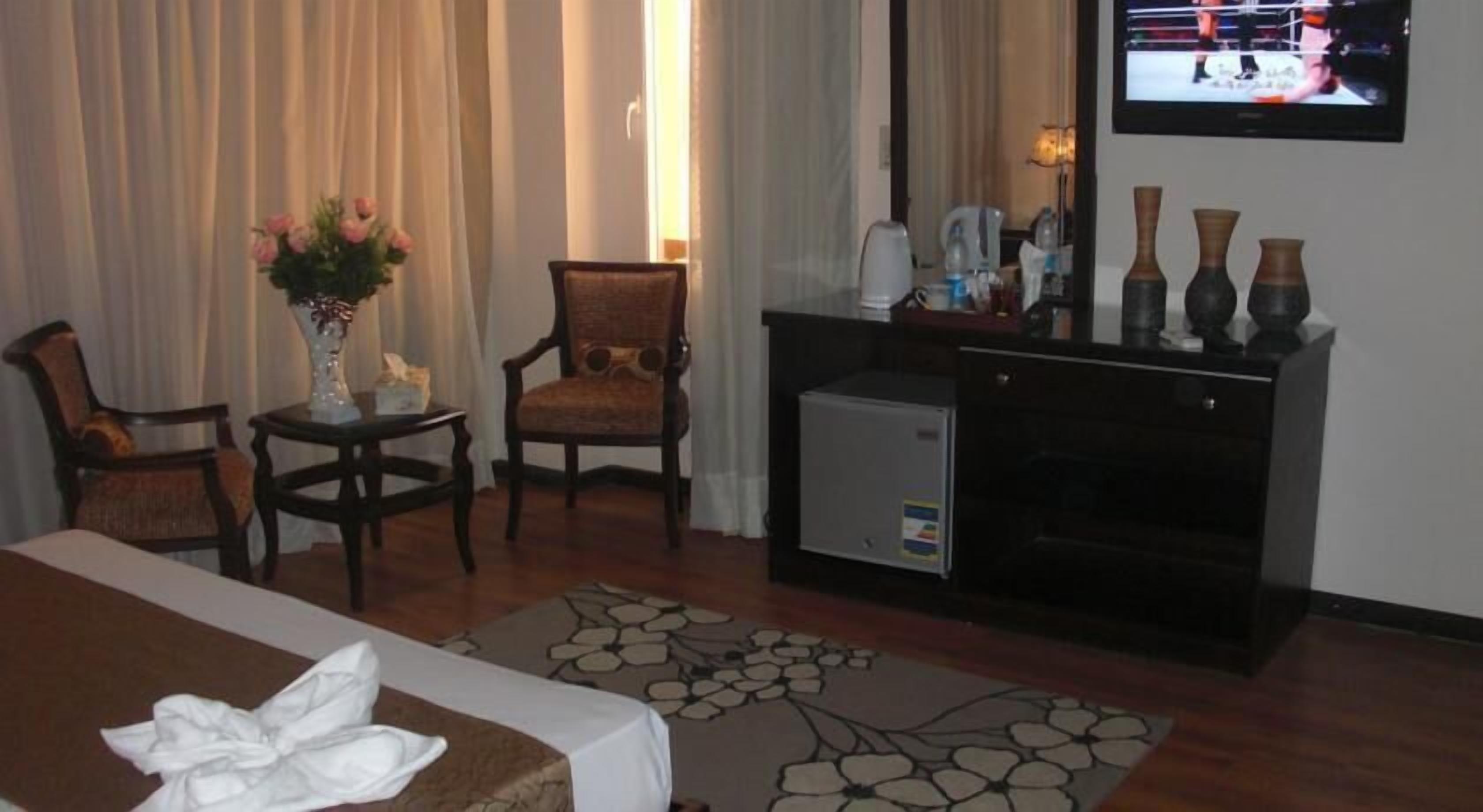 A hotel room with a bed, two chairs, a small table with a vase, a television, and a fireplace.